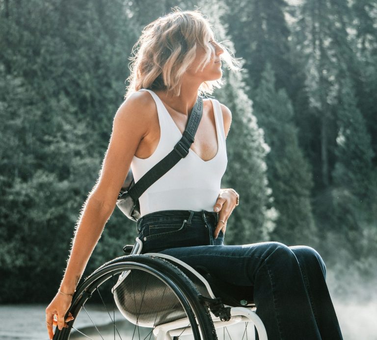 Blonde Female in Customized Wheelchair in front of dark green trees with sun shining on her face